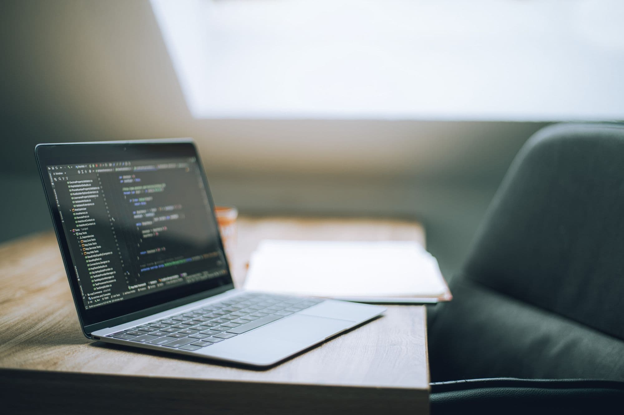 An open laptop showing a code editor on a wooden table beside a window.